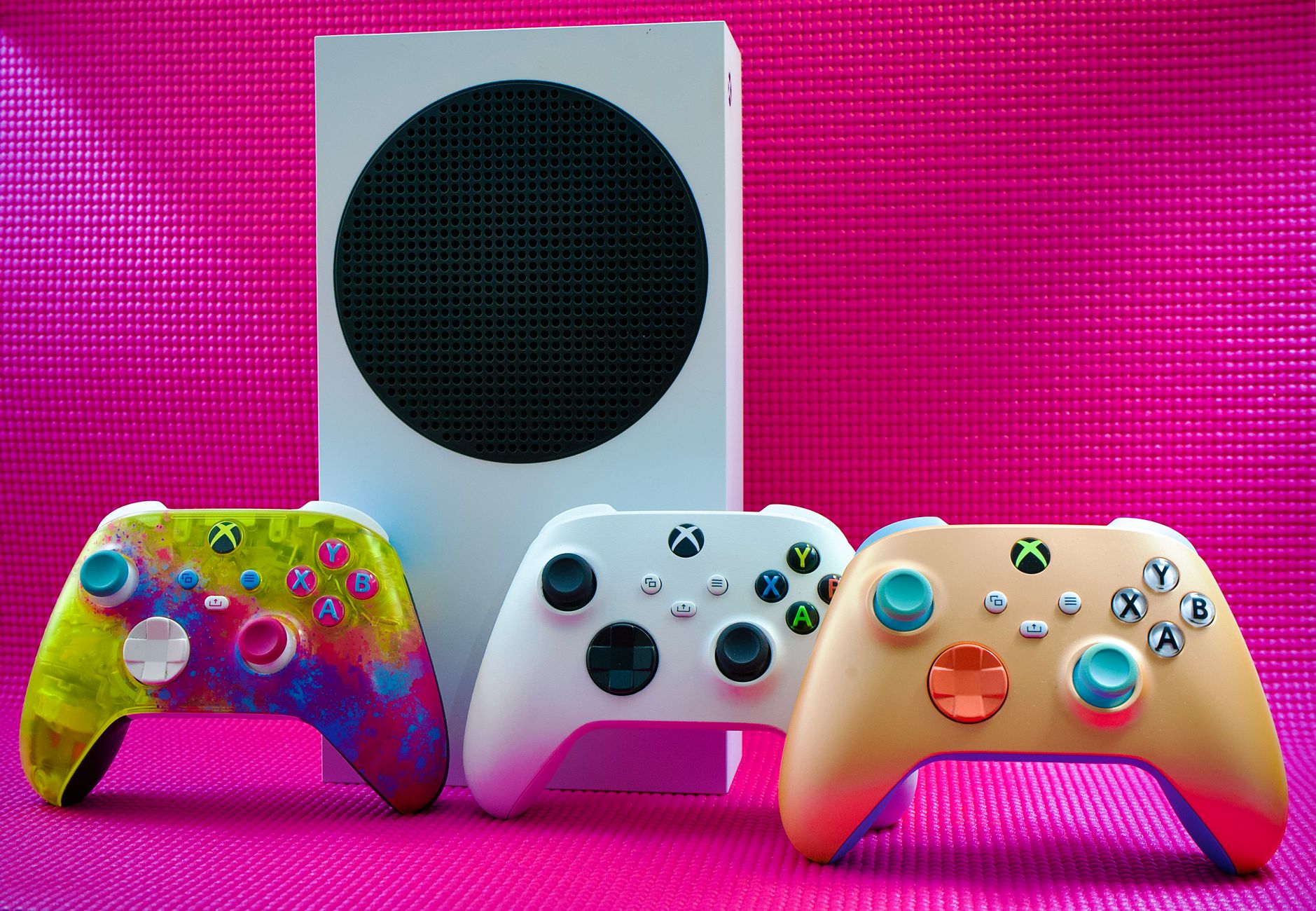 Vibrant gaming controllers displayed with a console on a bold pink backdrop.