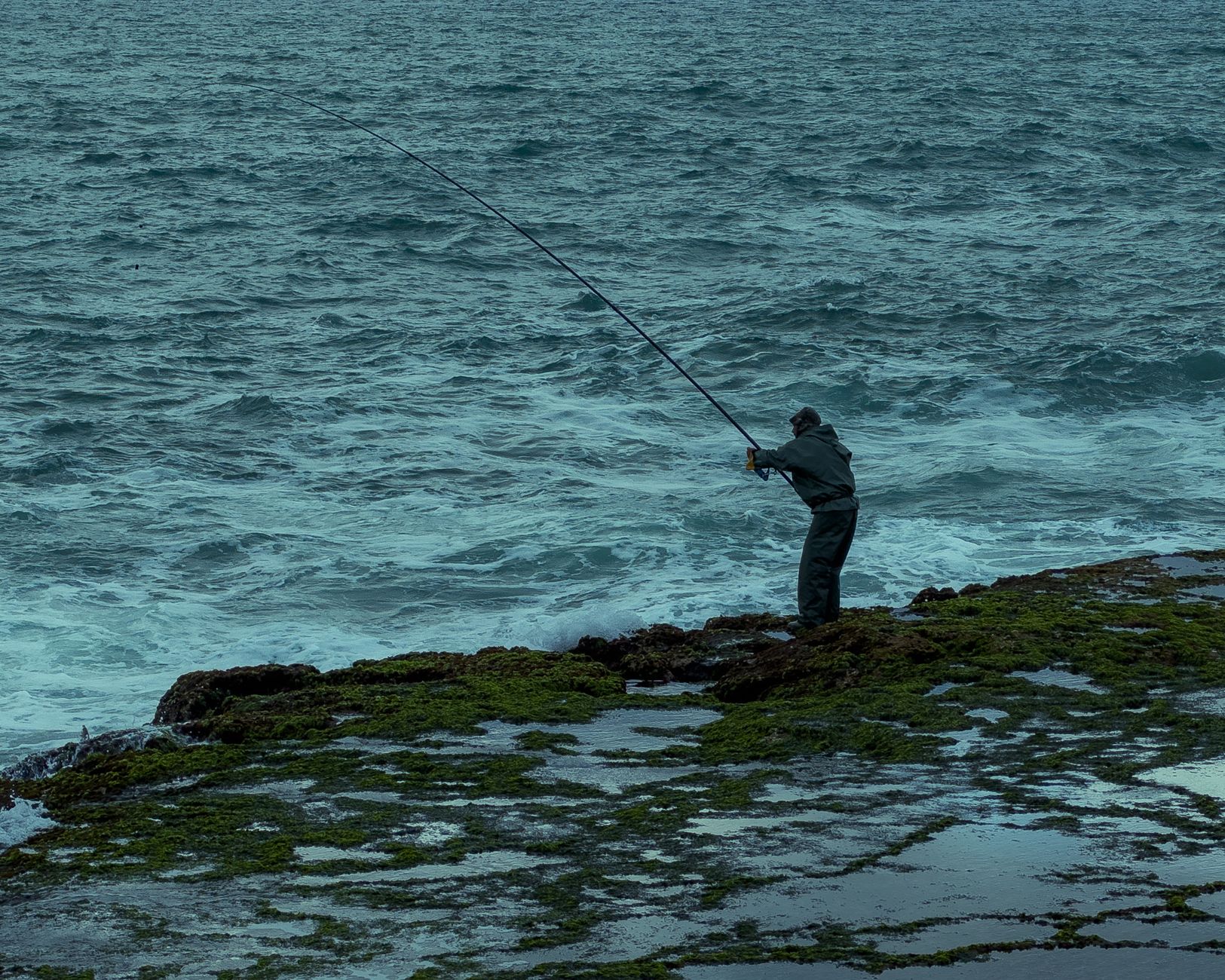A lone fisherman casts his line into the ocean on a rocky coastline.