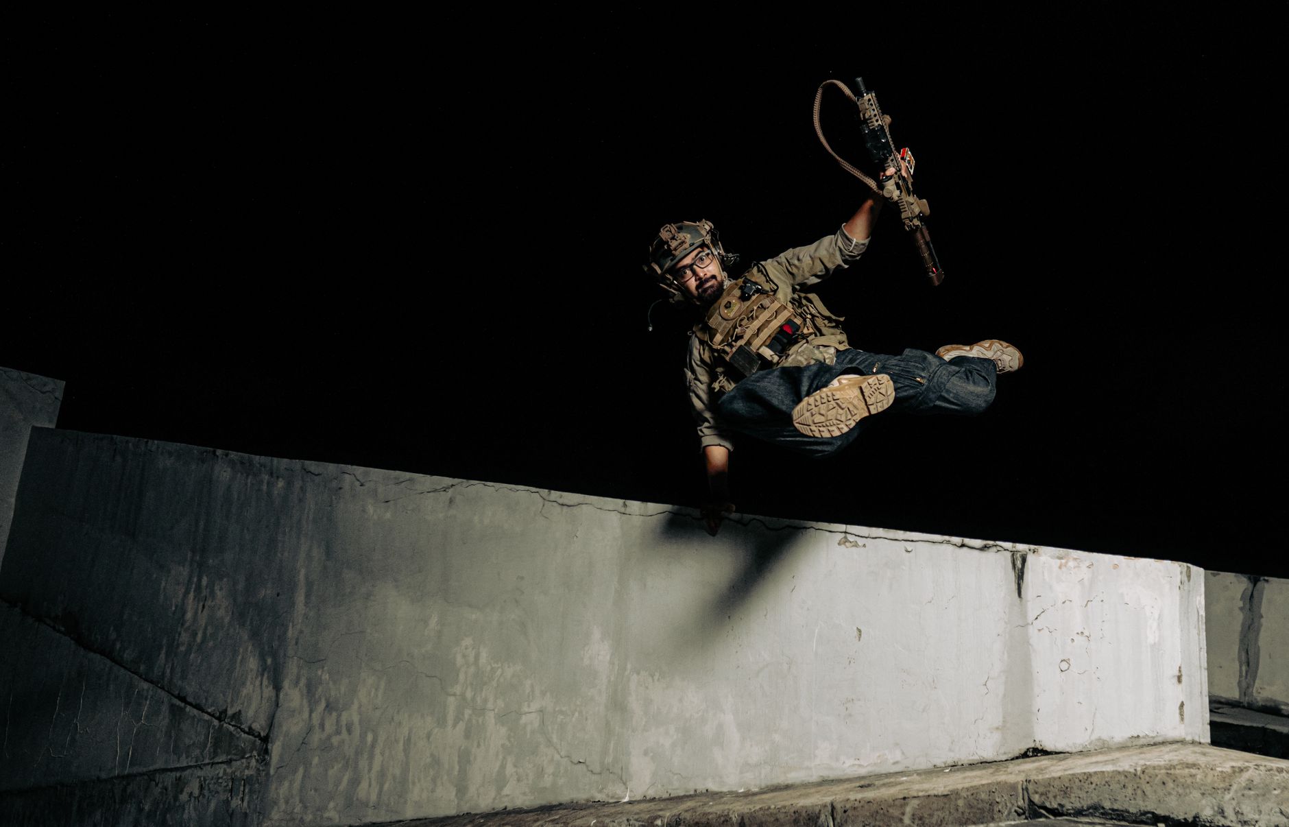 An action-packed shot of a soldier jumping midair over a wall with a weapon at night.