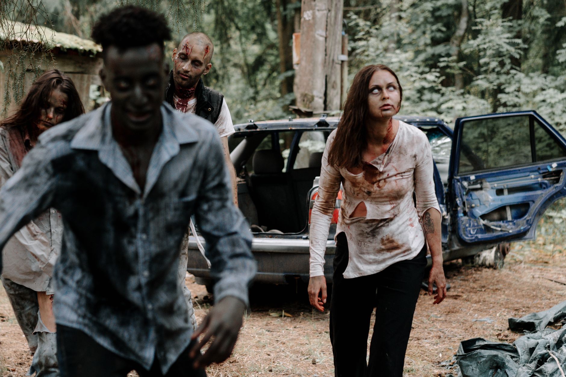 Group of realistic zombies in a forest setting with a wrecked car nearby.