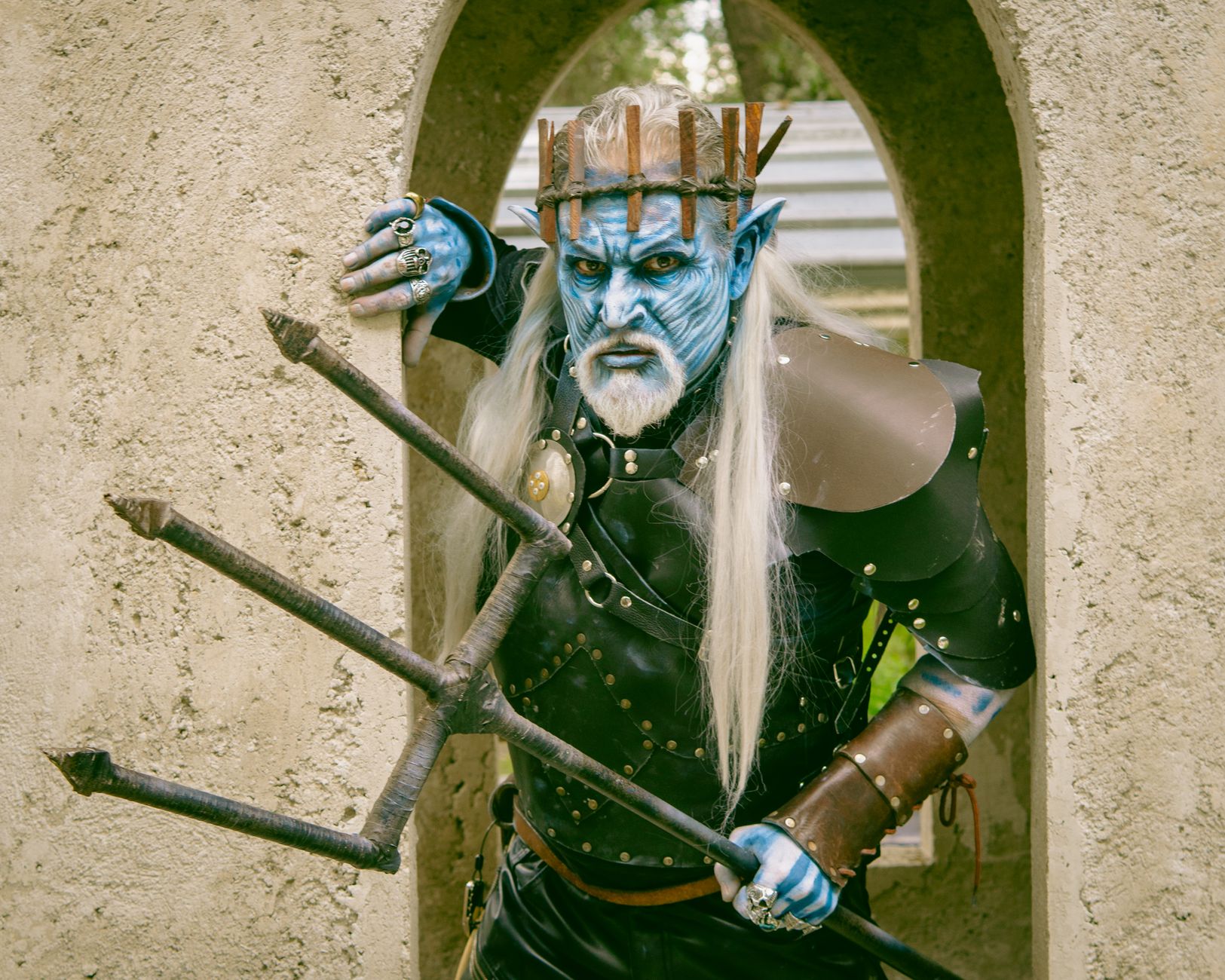 Blue-skinned demon character in leather armor posing with trident in a cemetery archway.