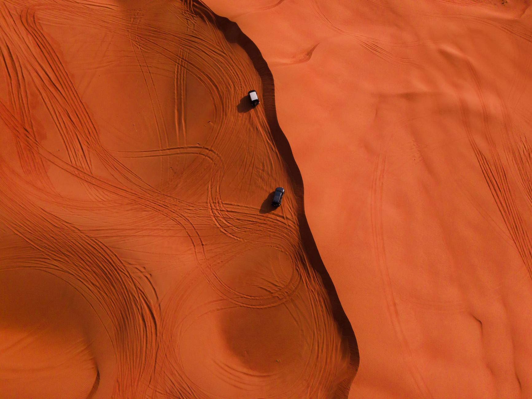 Stunning aerial shot of two cars making tracks on vibrant red sand dunes in a desert landscape.