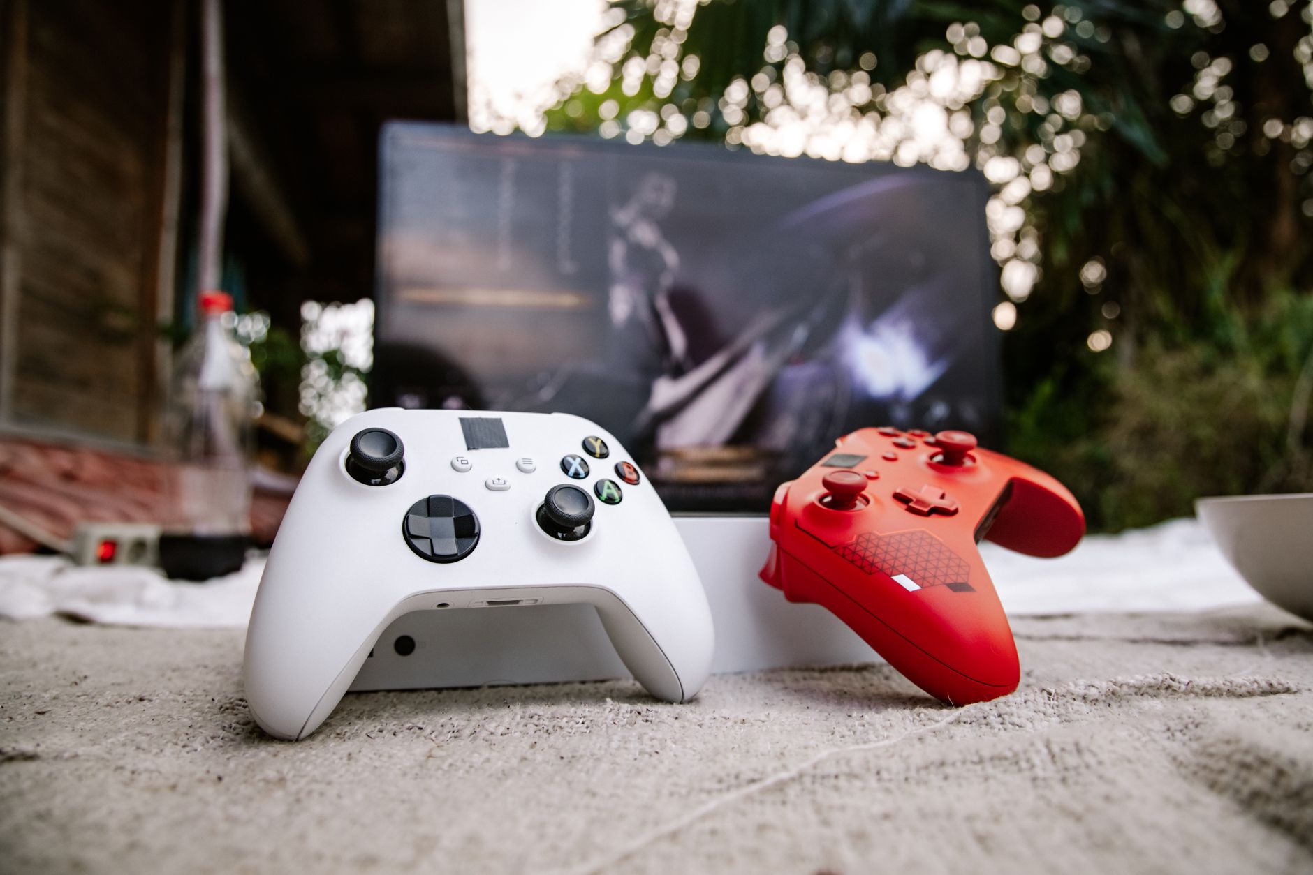 White and red Xbox wireless controllers set outdoors. A computer screen is blurred in the background.
