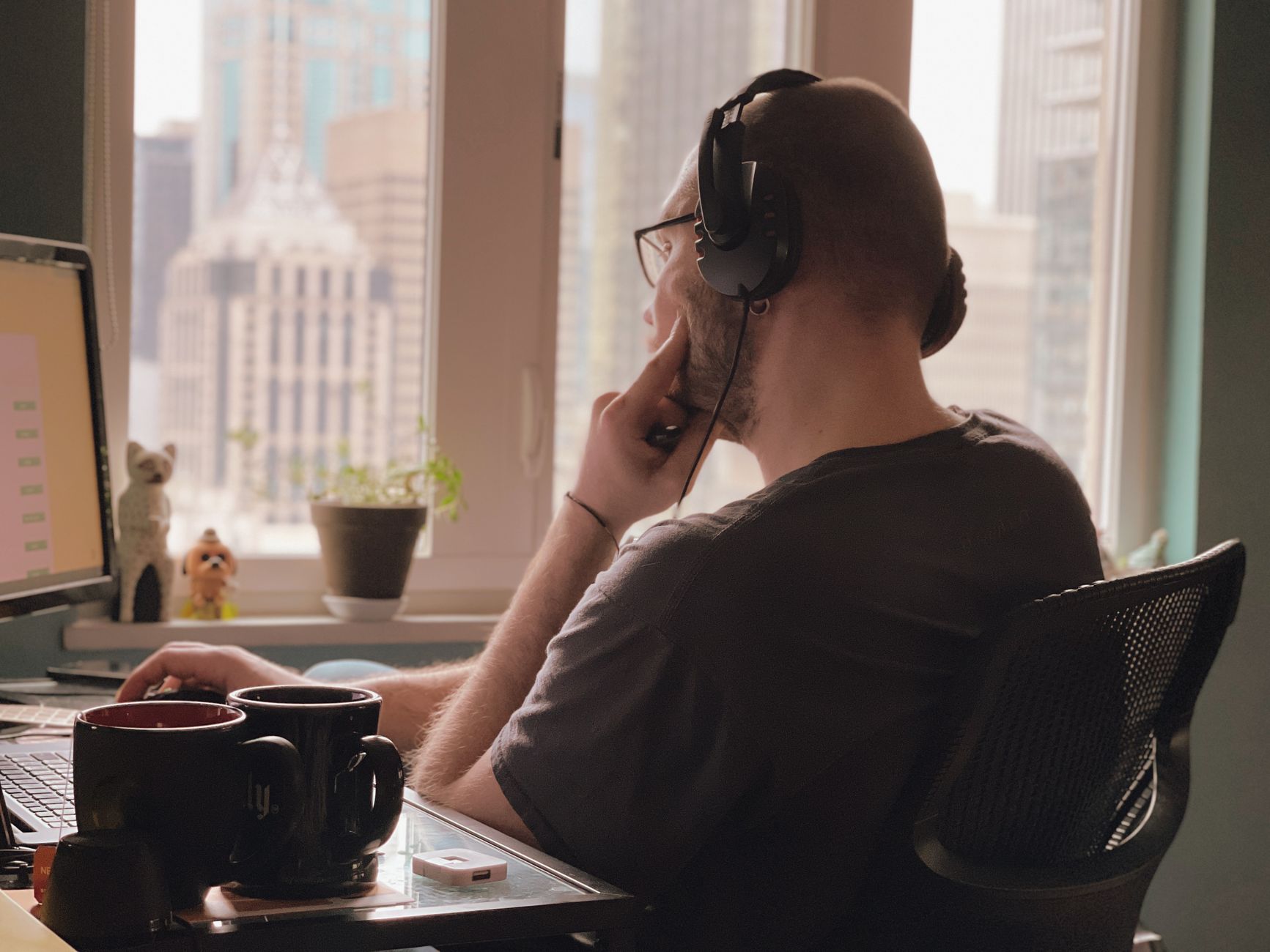 A man wearing headphones works at his desk overlooking a city skyline from a home office.