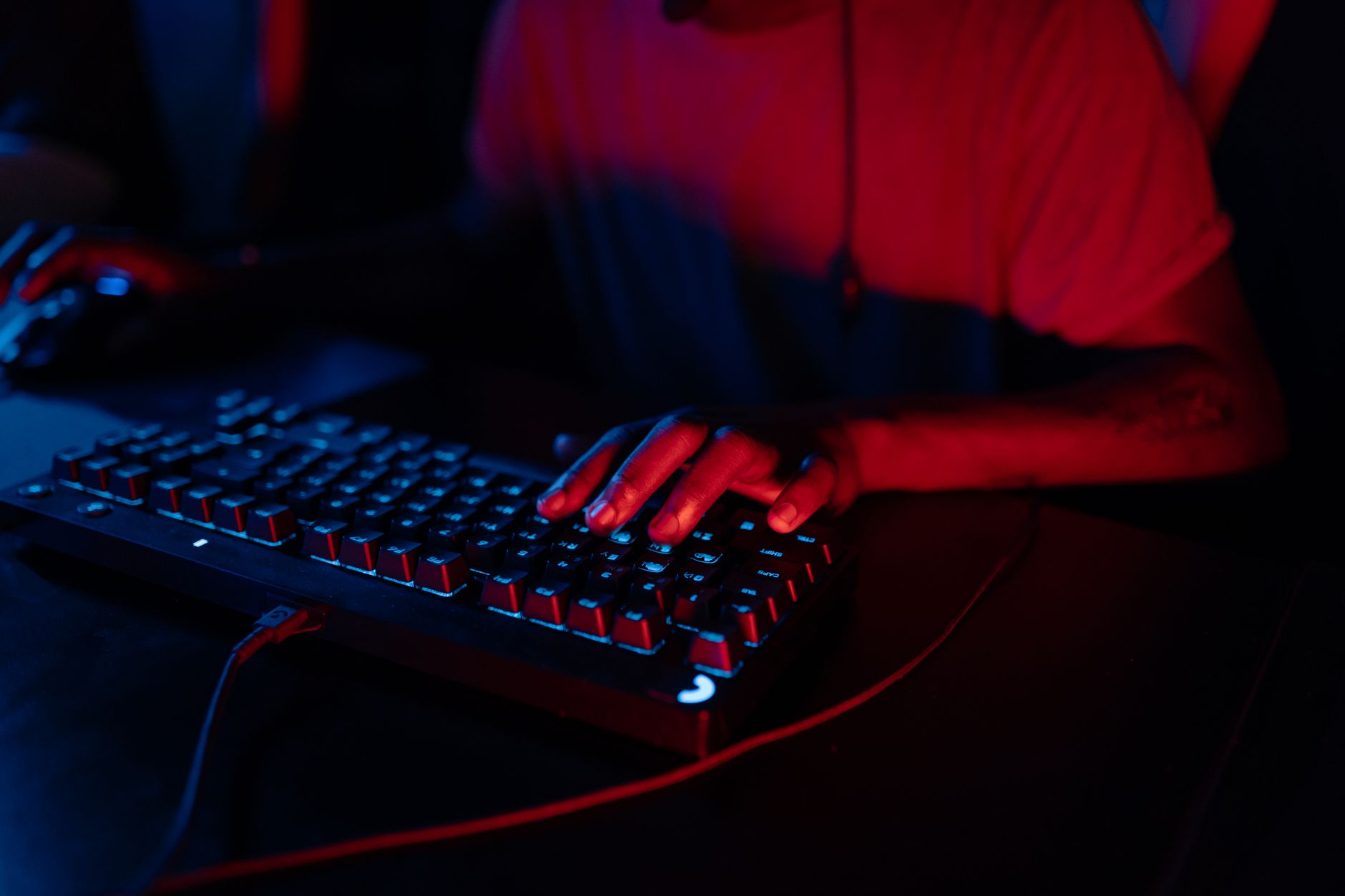Focus on hands typing on an RGB gaming keyboard under ambient red and blue lighting.