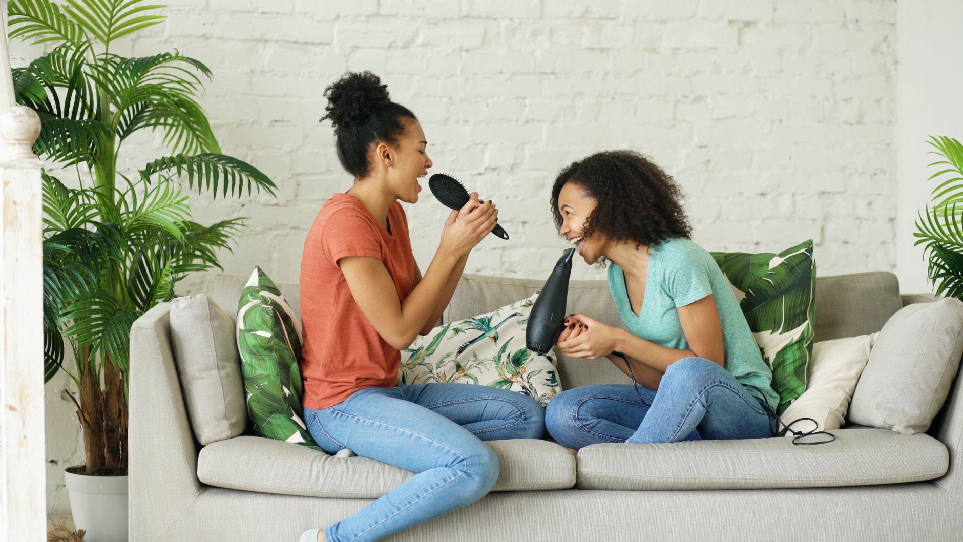 Two young women joyfully singing into hairbrush and hairdryer while sitting on a cozy couch in a bright living room.